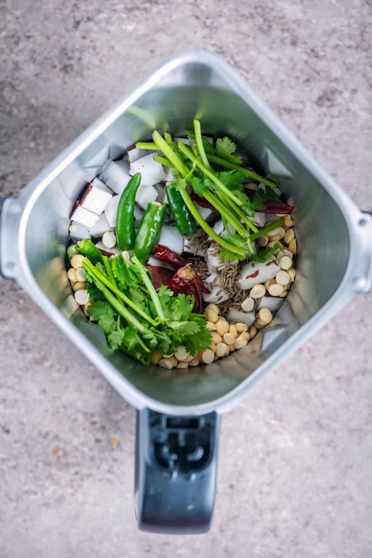 Grated coconut, dried red chillies, green chillies, cumin seeds, puttu kadalai, and cilantro stalks placed in a blender jar for white pumpkin curry masala.