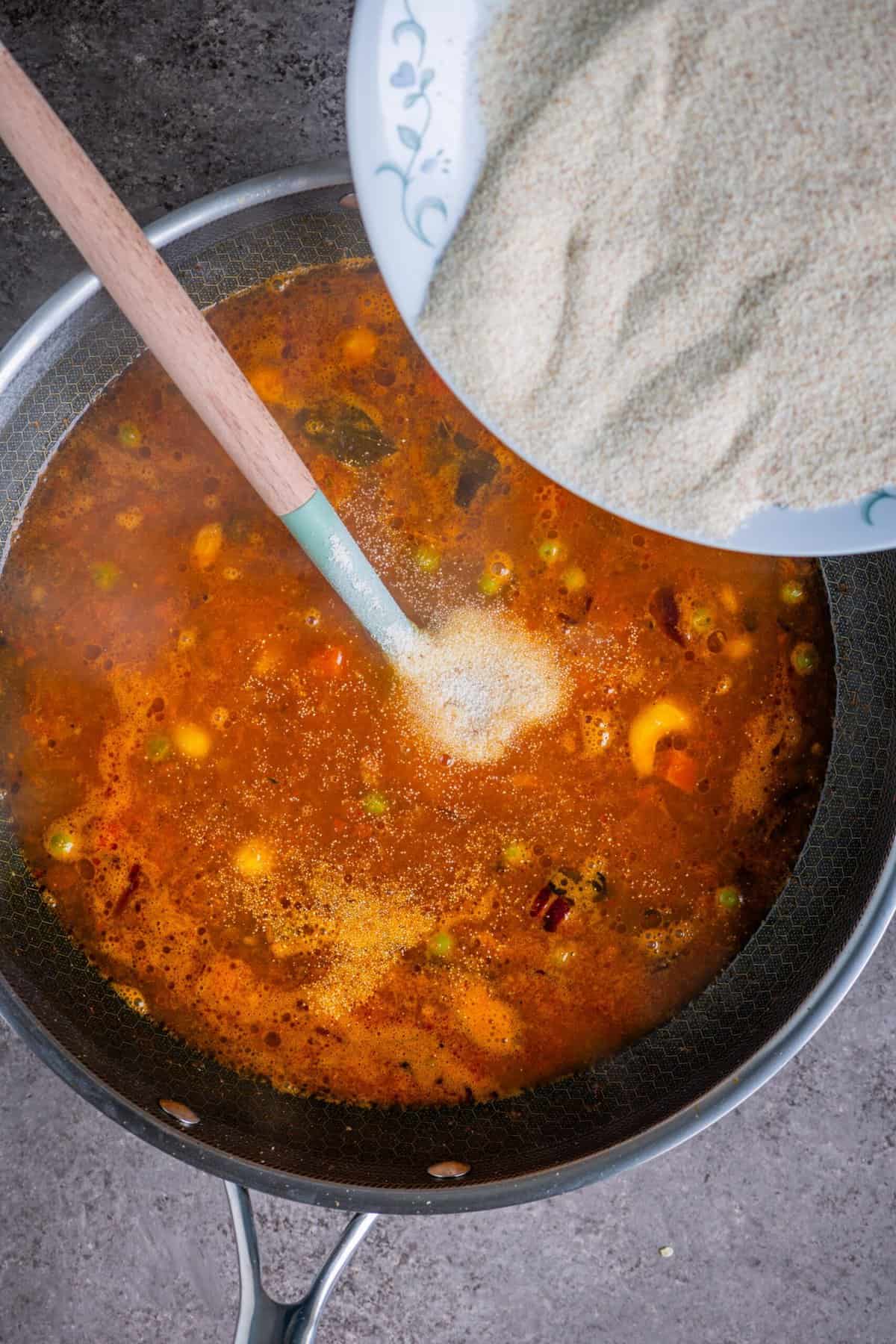 Roasted rava being added gradually to tomato mixture while stirring for upma.