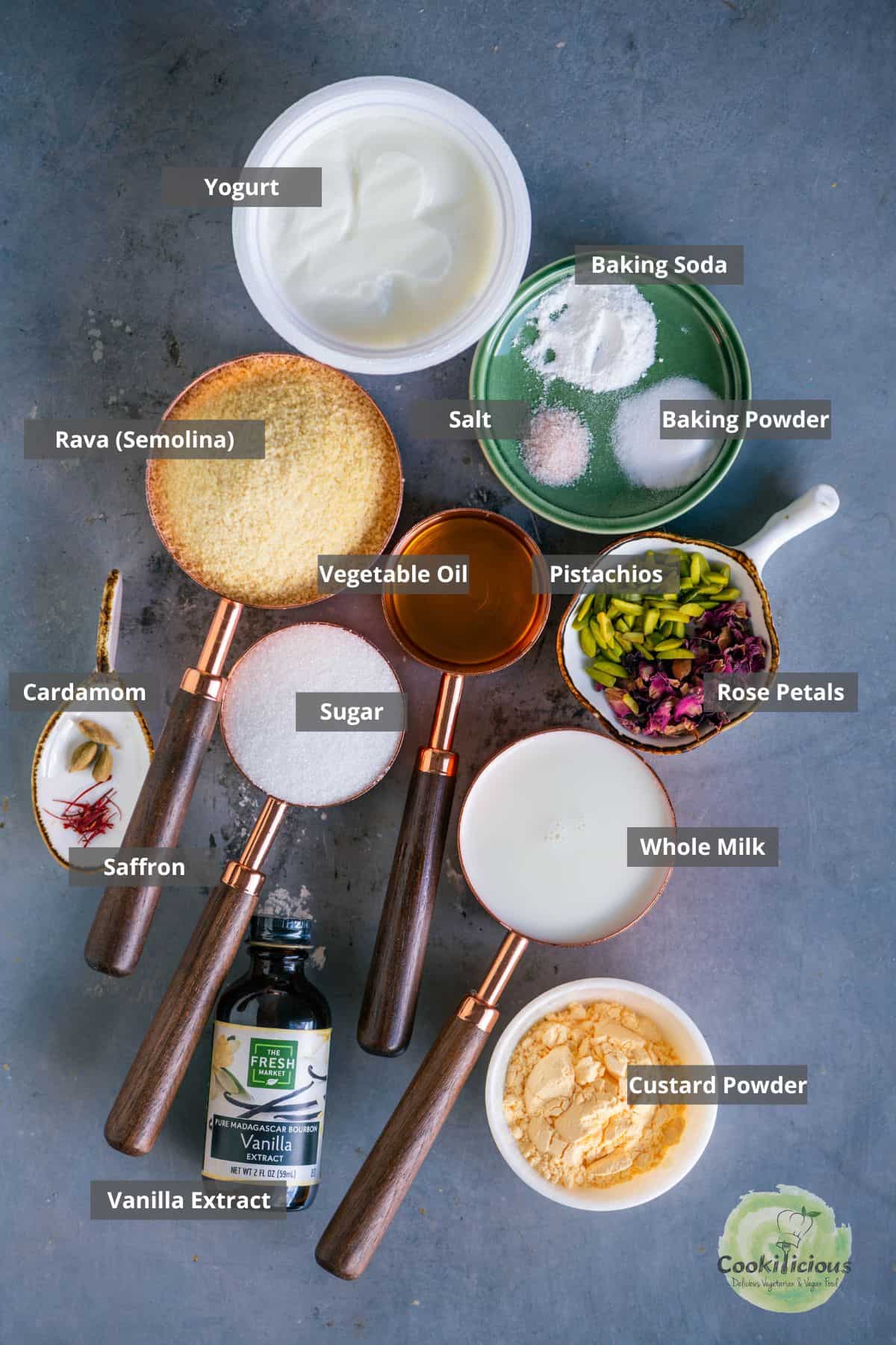 Ingredients for eggless Gulab Jamun Cupcakes — rava, yogurt, milk, custard powder, sugar, cardamom, nuts, vanilla essence, and saffron placed on a table with labels on them.