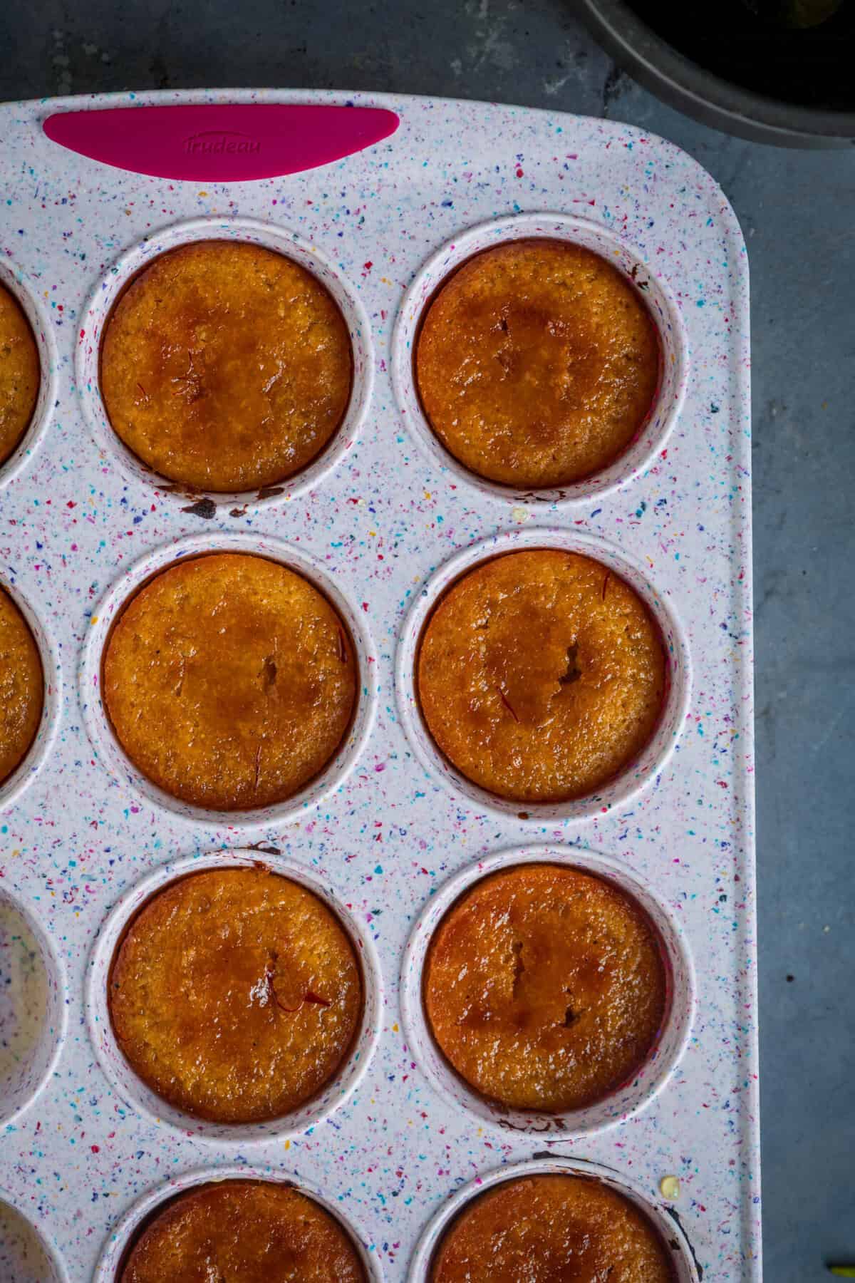Pouring warm cardamom sugar syrup over baked gulab jamun cupcakes.