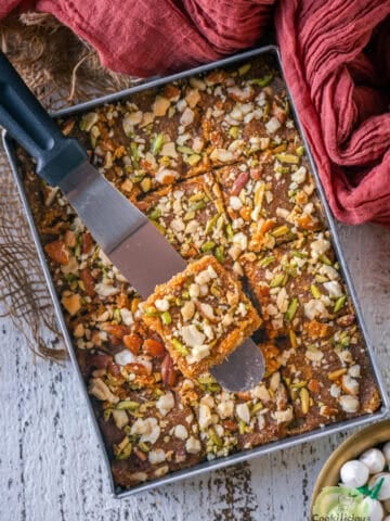 Caramel coconut barfi (nariyal barfi) being sliced into squares in the pan with a metal spatula.
