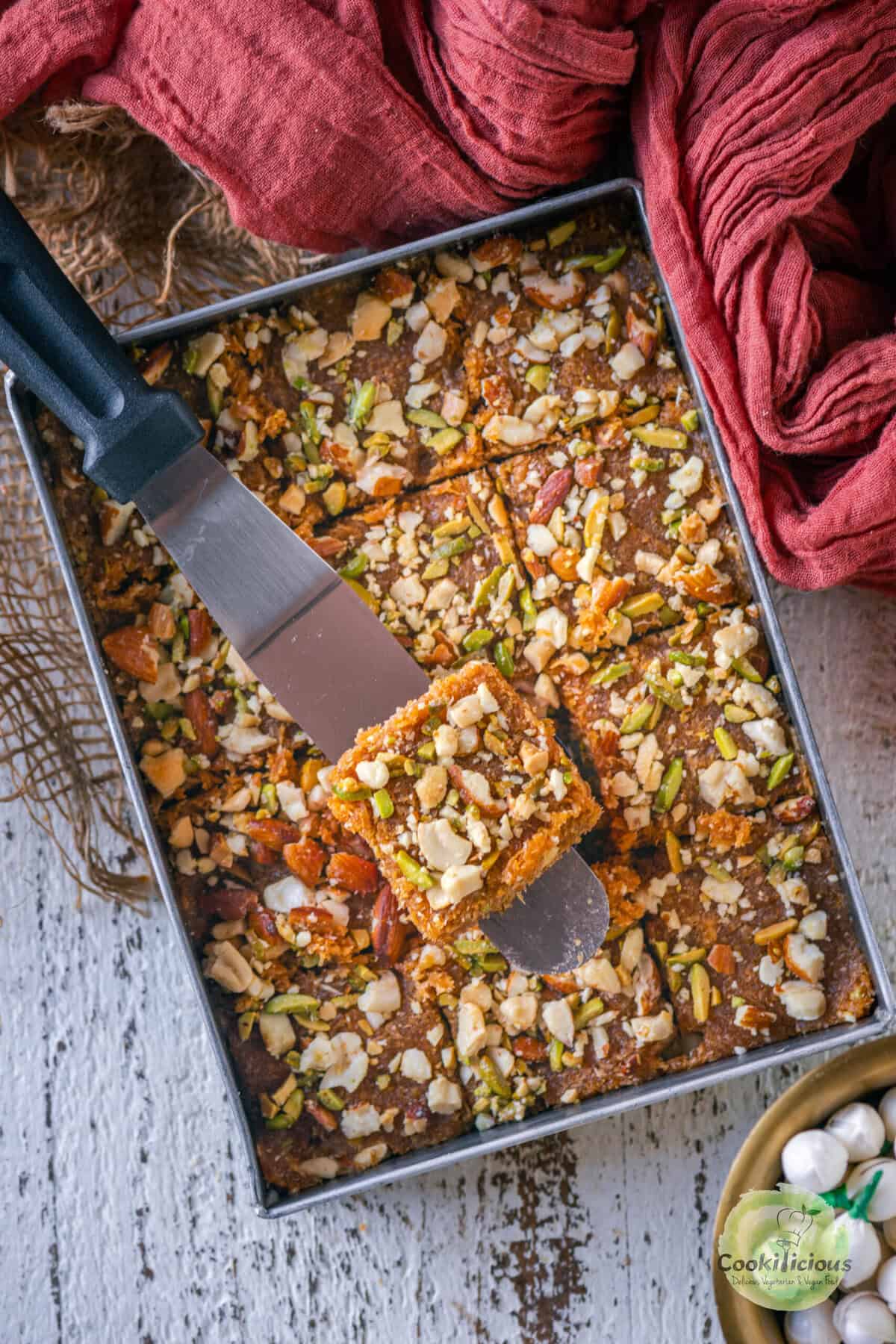 Caramel coconut barfi (nariyal barfi) being sliced into squares in the pan with a metal spatula.