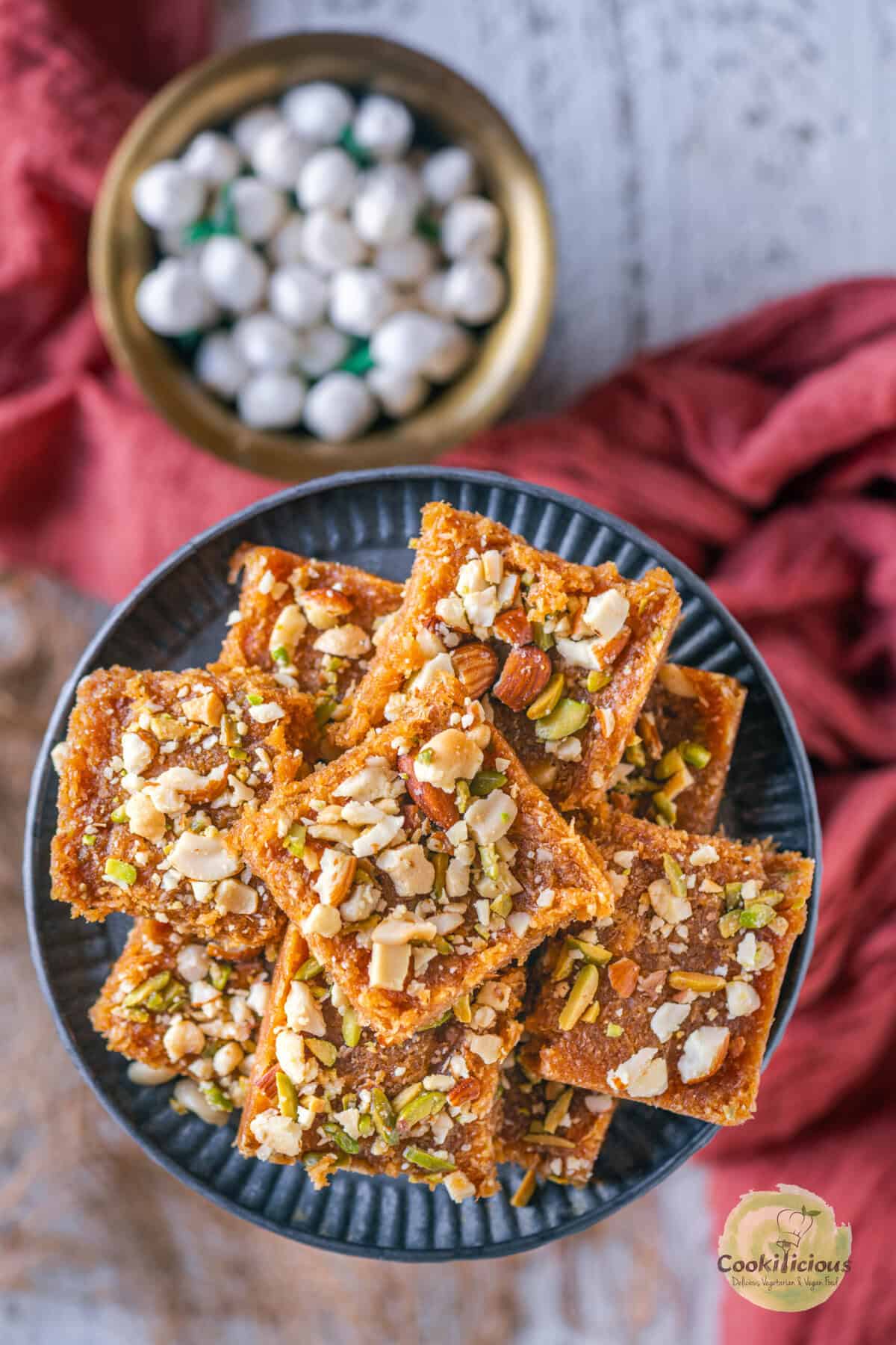 Stack of caramel coconut barfi pieces garnished with mixed nuts, served on a black plate.
